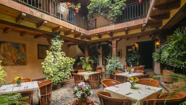 Un patio con varias mesas y sillas, rodeado de plantas en macetas y una escalera que lleva a un nivel superior.