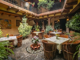 Un patio con varias mesas y sillas, rodeado de plantas en macetas y una escalera que lleva a un nivel superior.