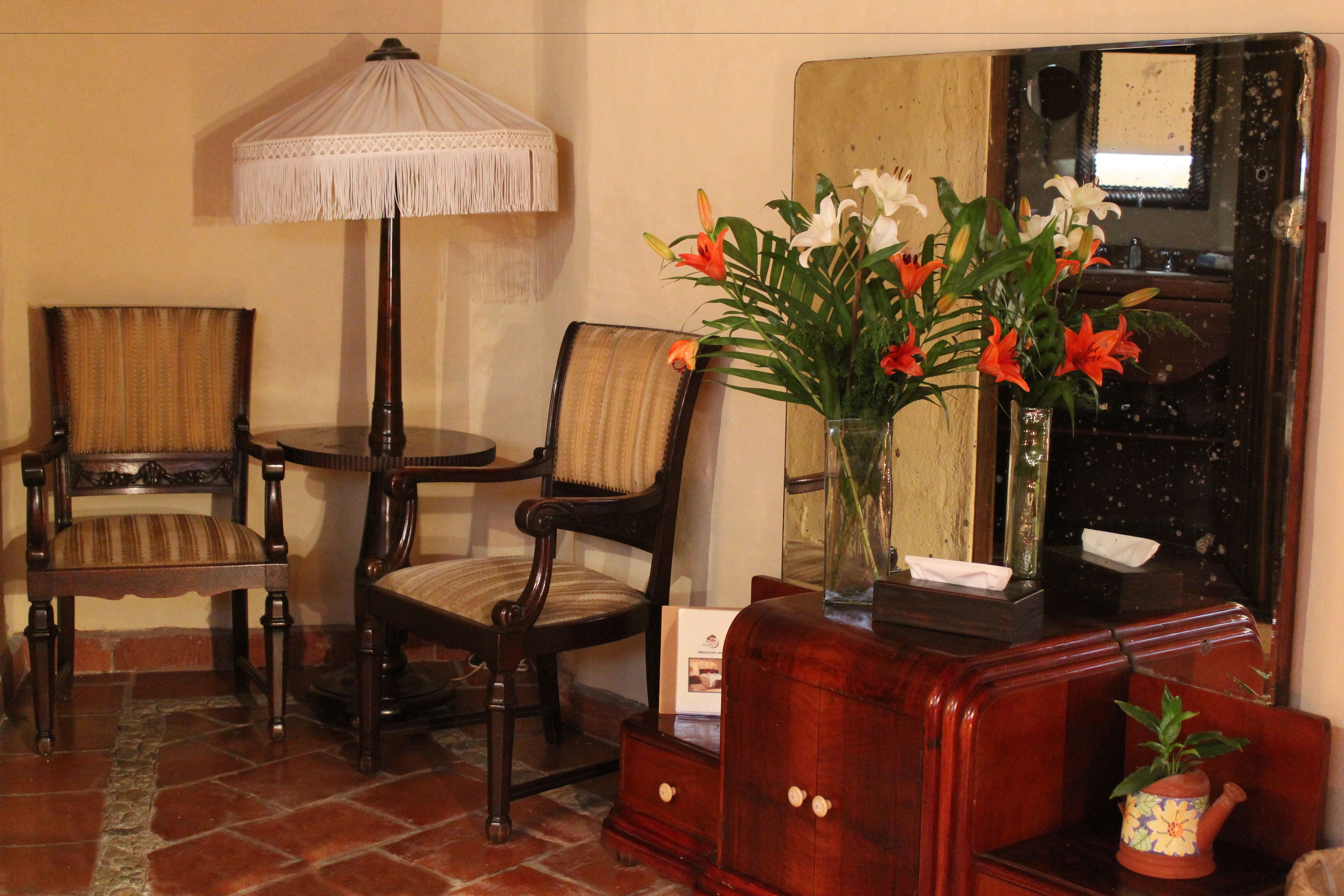 Una habitación con dos sillas de madera, una pequeña mesa redonda, una lámpara de pie, un espejo y un armario de madera con un jarrón de flores.