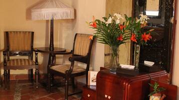 Una habitación con dos sillas de madera, una pequeña mesa redonda, una lámpara de pie, un espejo y un armario de madera con un jarrón de flores.