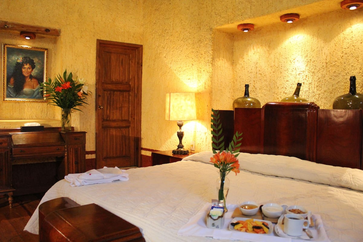 A hotel room with a bed, a desk, a lamp, a painting, and a tray with food and drinks.