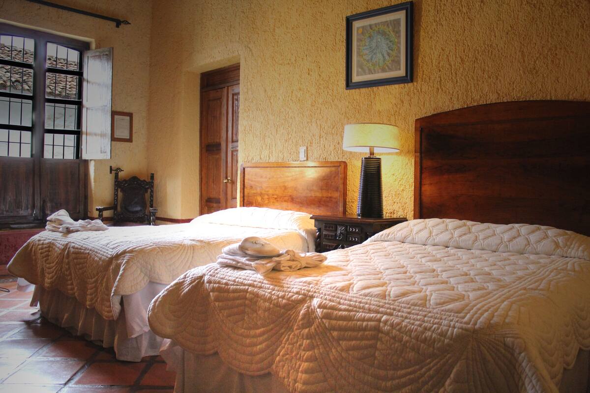 A hotel room with two beds, a wooden headboard, a nightstand with a lamp, and a door leading to another room.