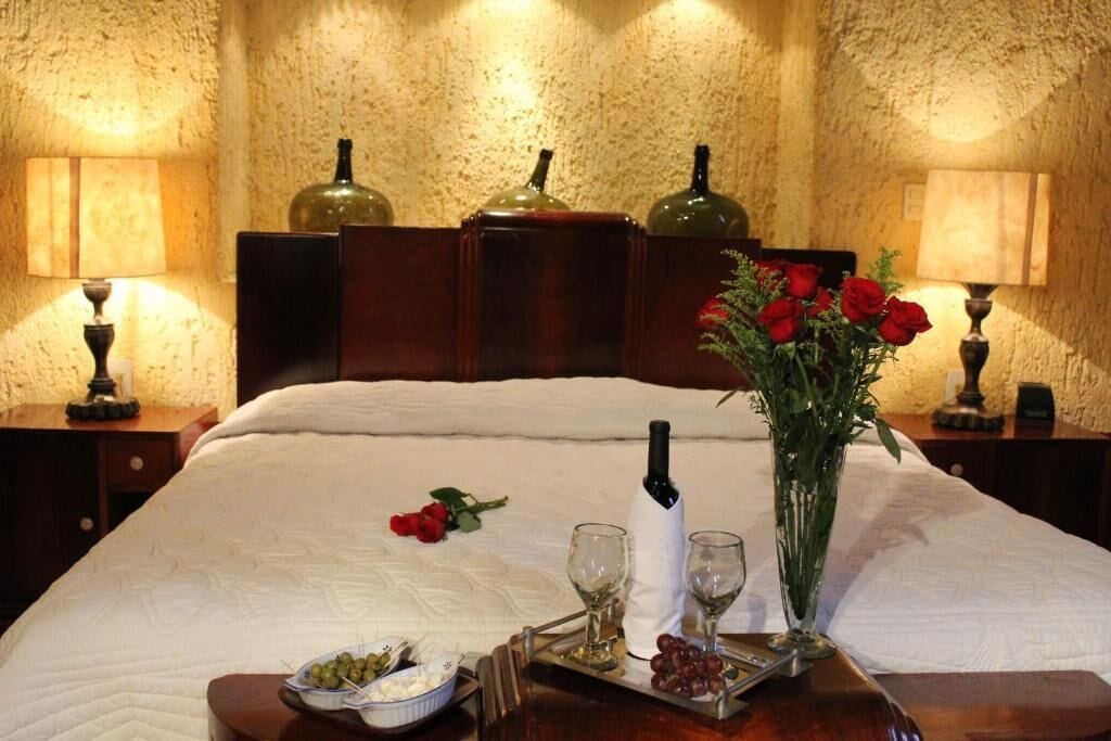 Un lit bien fait, recouvert d’une courtepointe blanche, une table avec des verres à vin et des raisins, et un vase de roses rouges posé sur une table de chevet en bois.