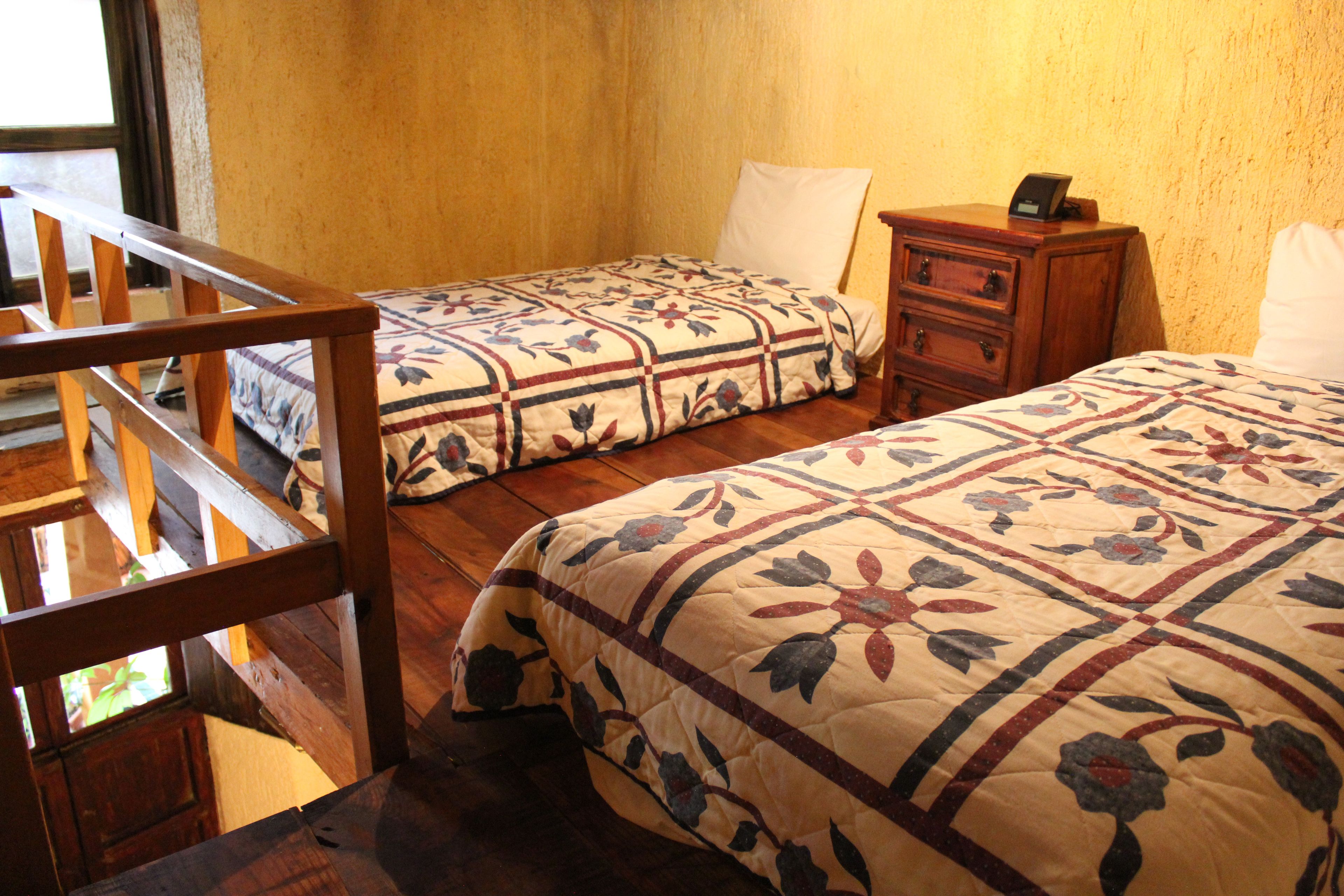 Una habitación con dos camas, una silla de madera, una mesita de noche con un reloj y una ventana que da al exterior.