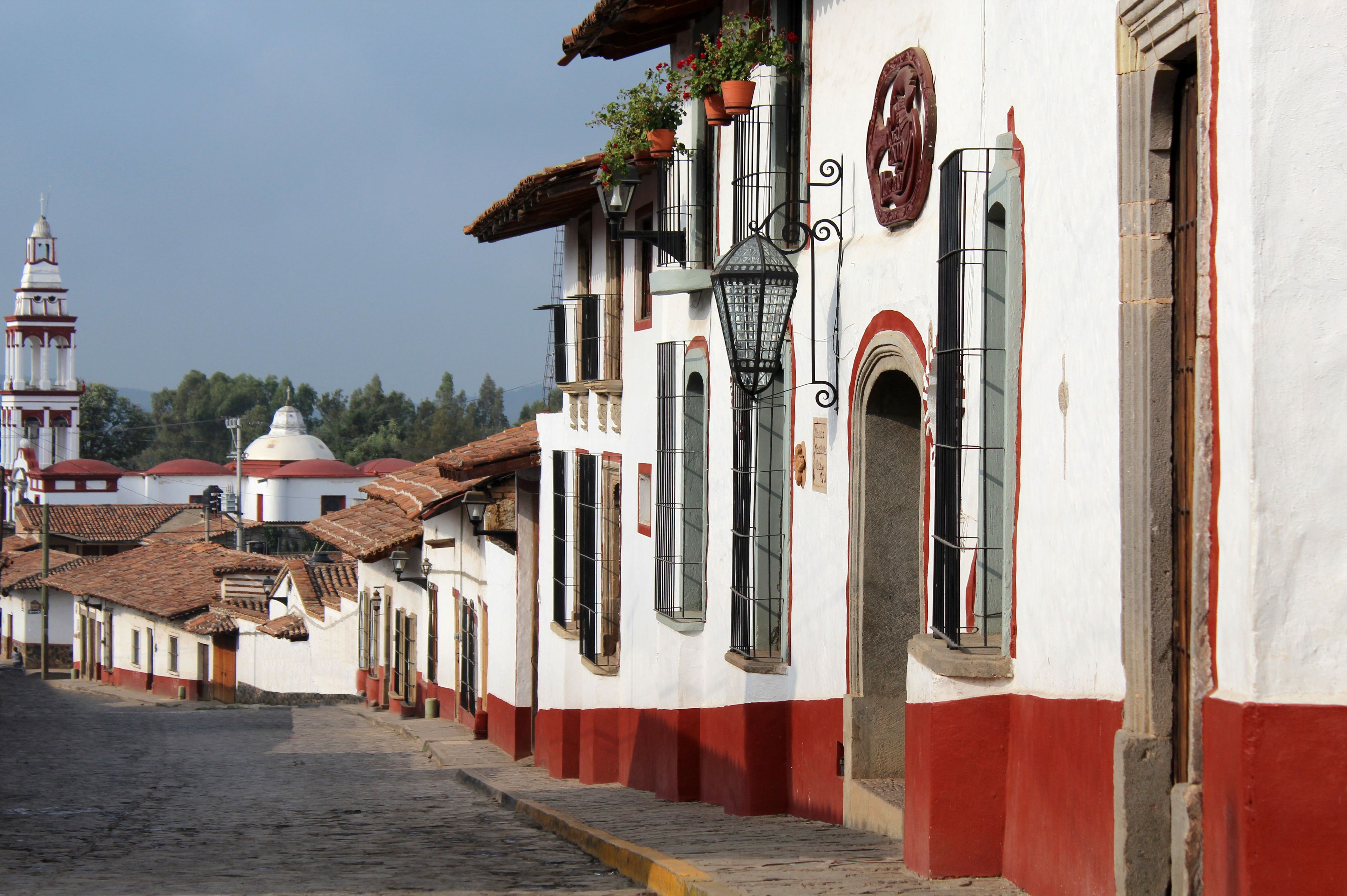 A cobblestone street lined with traditional buildings featuring red and white facades, a bell tower, and a church dome in the background.
