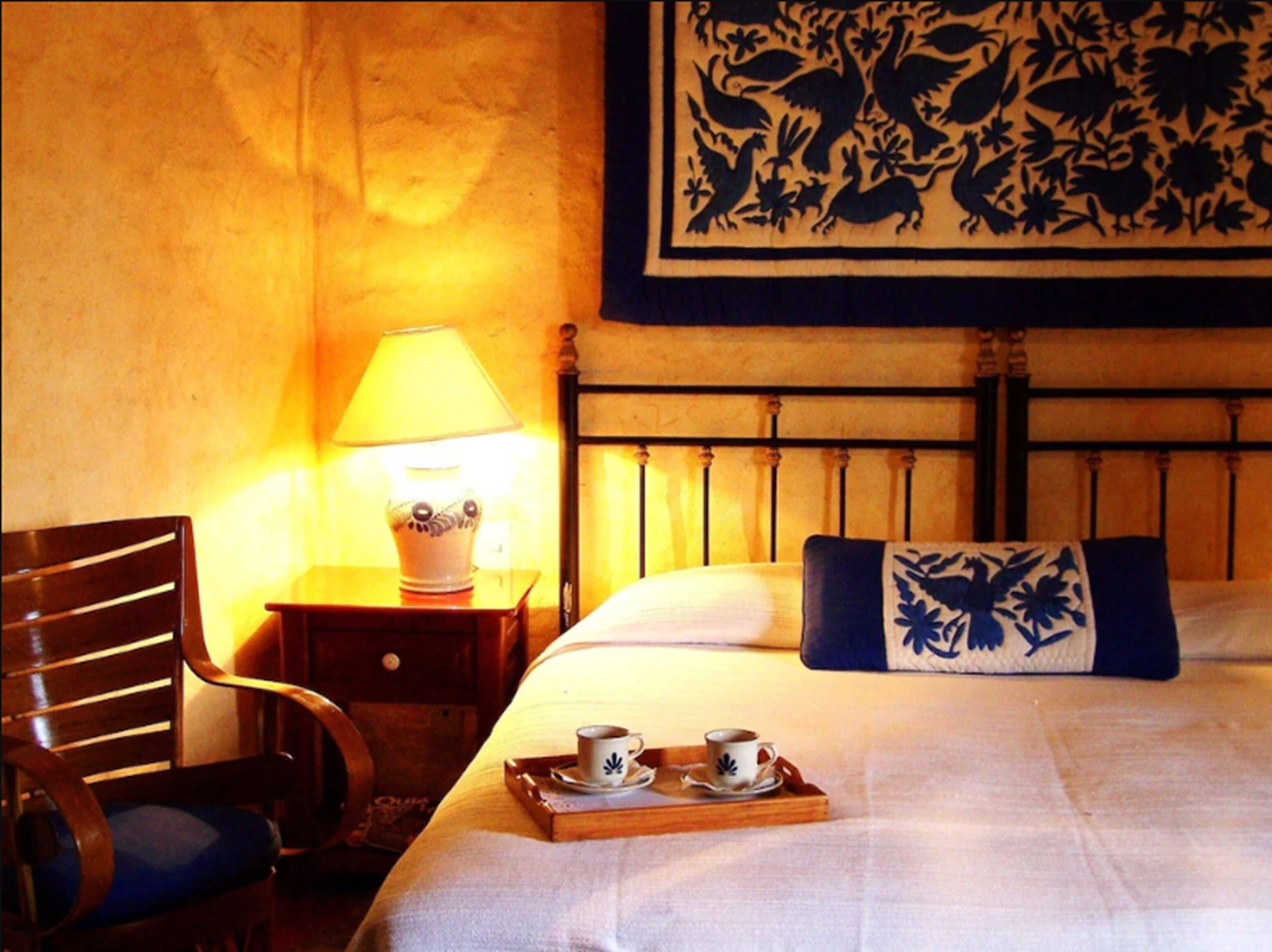 Habitación de hotel con una cama, una silla, una mesita de noche con una lámpara y una bandeja con dos tazas.