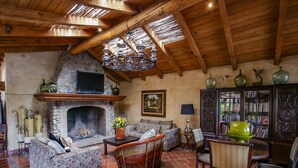 Una sala de estar rústica con chimenea de piedra, vigas de madera y pared de piedra. Hay un sofá, una silla y una mesa con un jarrón.