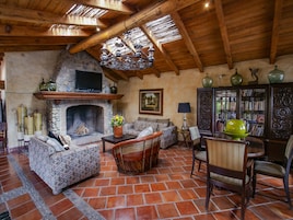 Una sala de estar rústica con chimenea de piedra, vigas de madera y pared de piedra. Hay un sofá, una silla y una mesa con un jarrón.