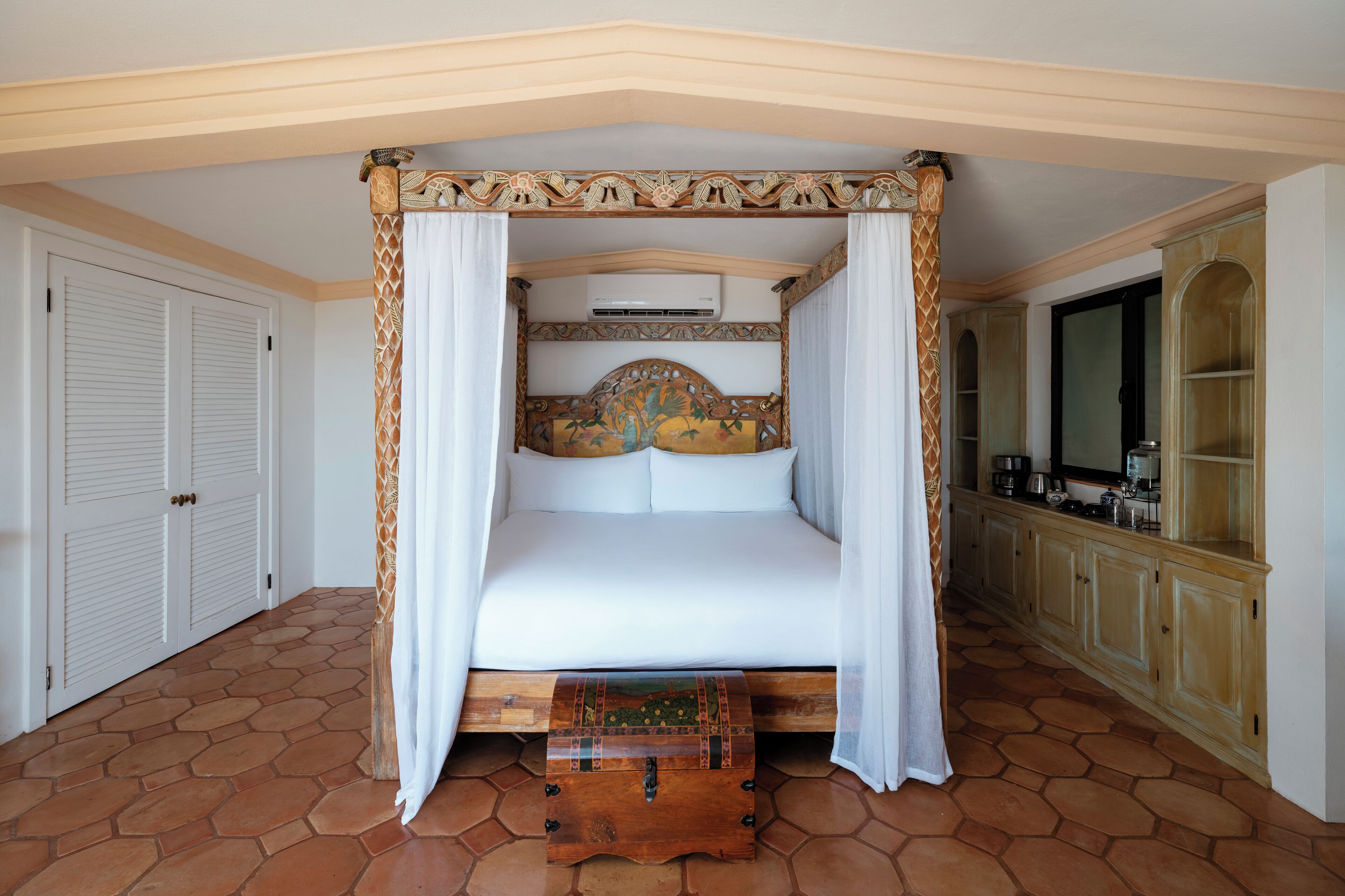 A four-poster bed with a decorative headboard, a wooden chest at the foot of the bed, and a kitchen area with cabinets and a television.