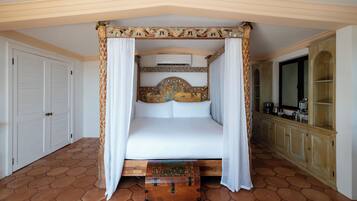 A four-poster bed with a decorative headboard, a wooden chest at the foot of the bed, and a kitchen area with cabinets and a television.