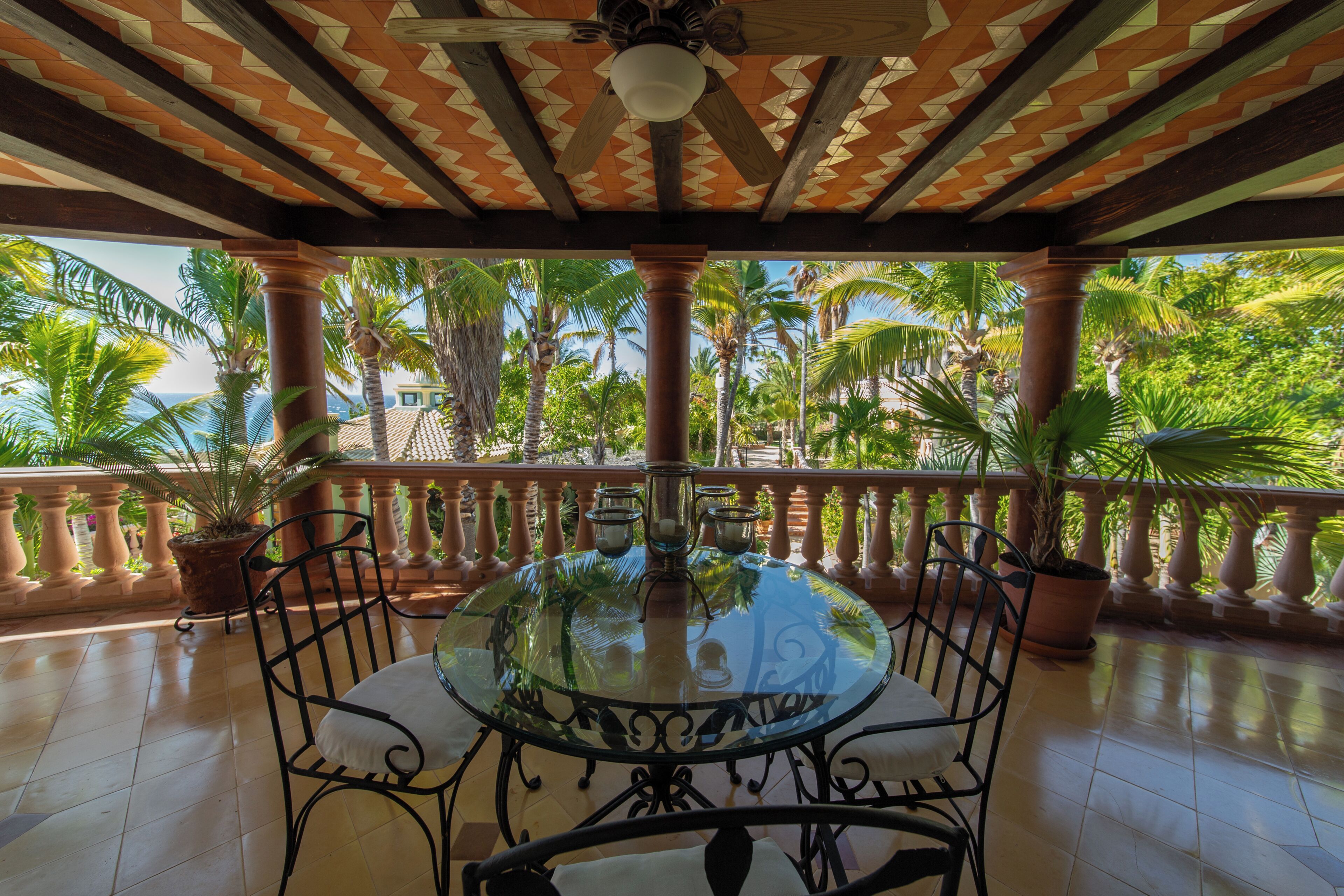 A covered outdoor area with a glass table and chairs, surrounded by potted plants and palm trees.