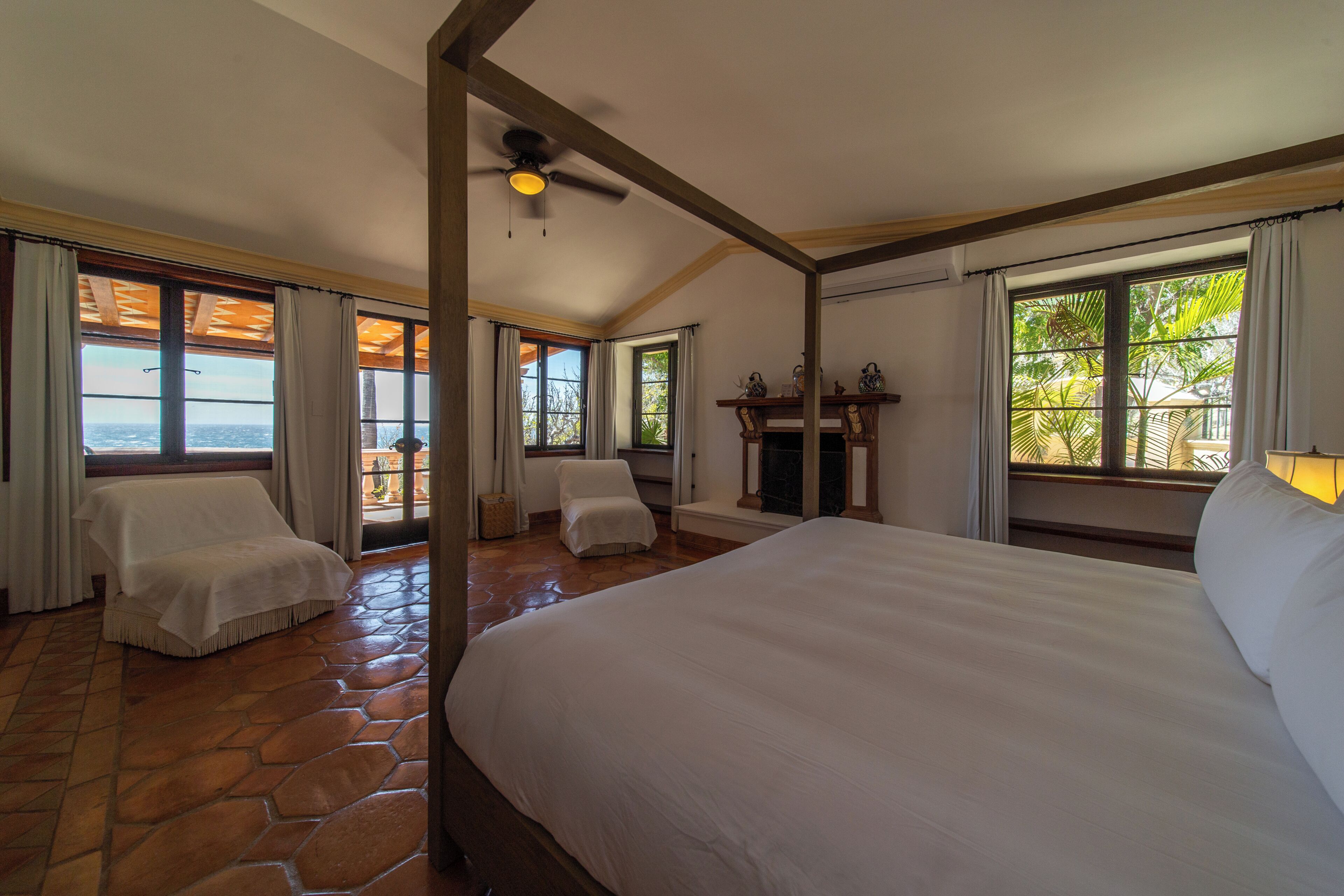 A bedroom with a large bed, two chairs, a fireplace, and large windows with curtains.