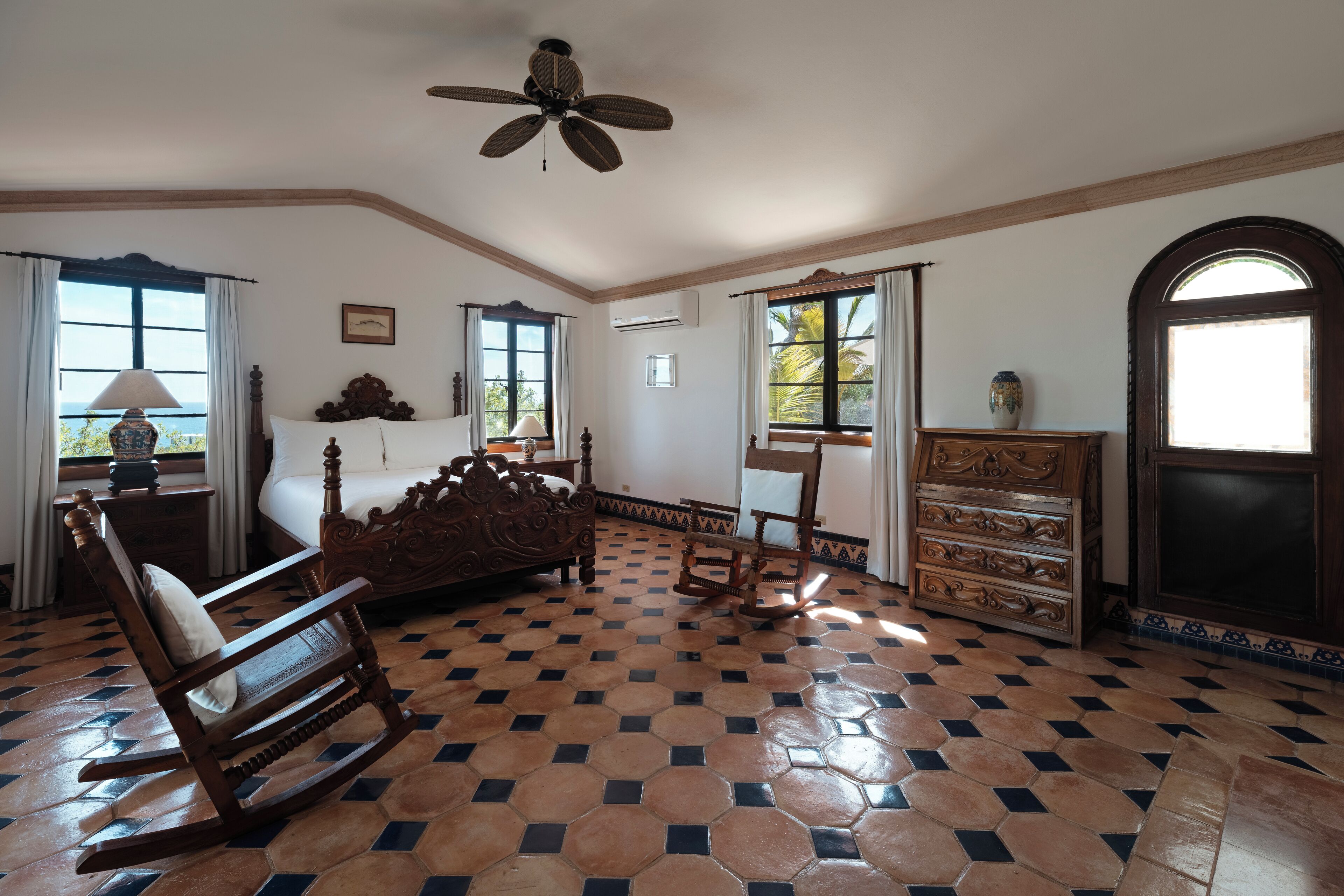 A room with a checkered floor, a rocking chair, a bed with a headboard, a wooden dresser, and a ceiling fan.