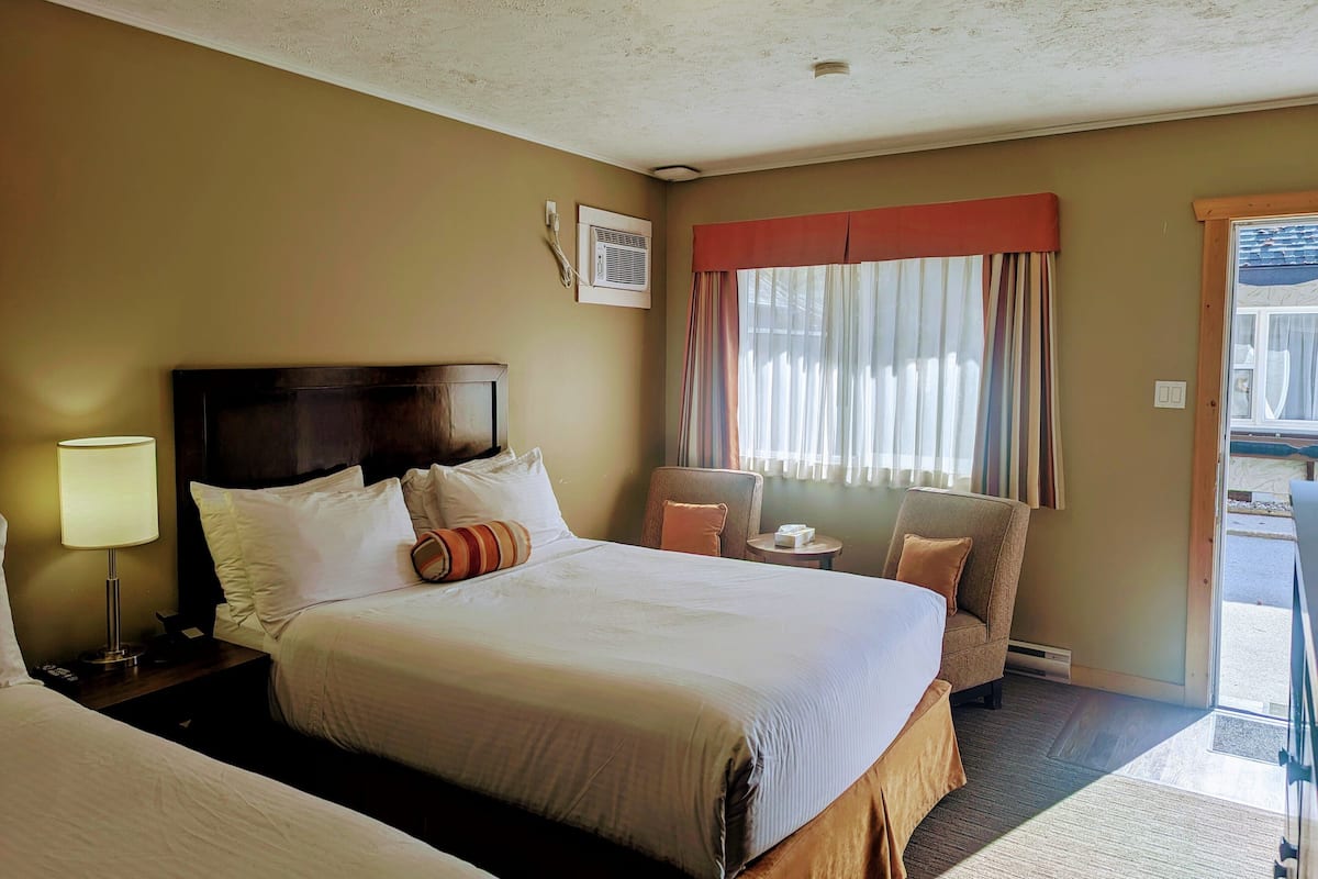 A hotel room with a large bed, a bedside table with a lamp, a chair, a window with curtains, and a view of the outside.