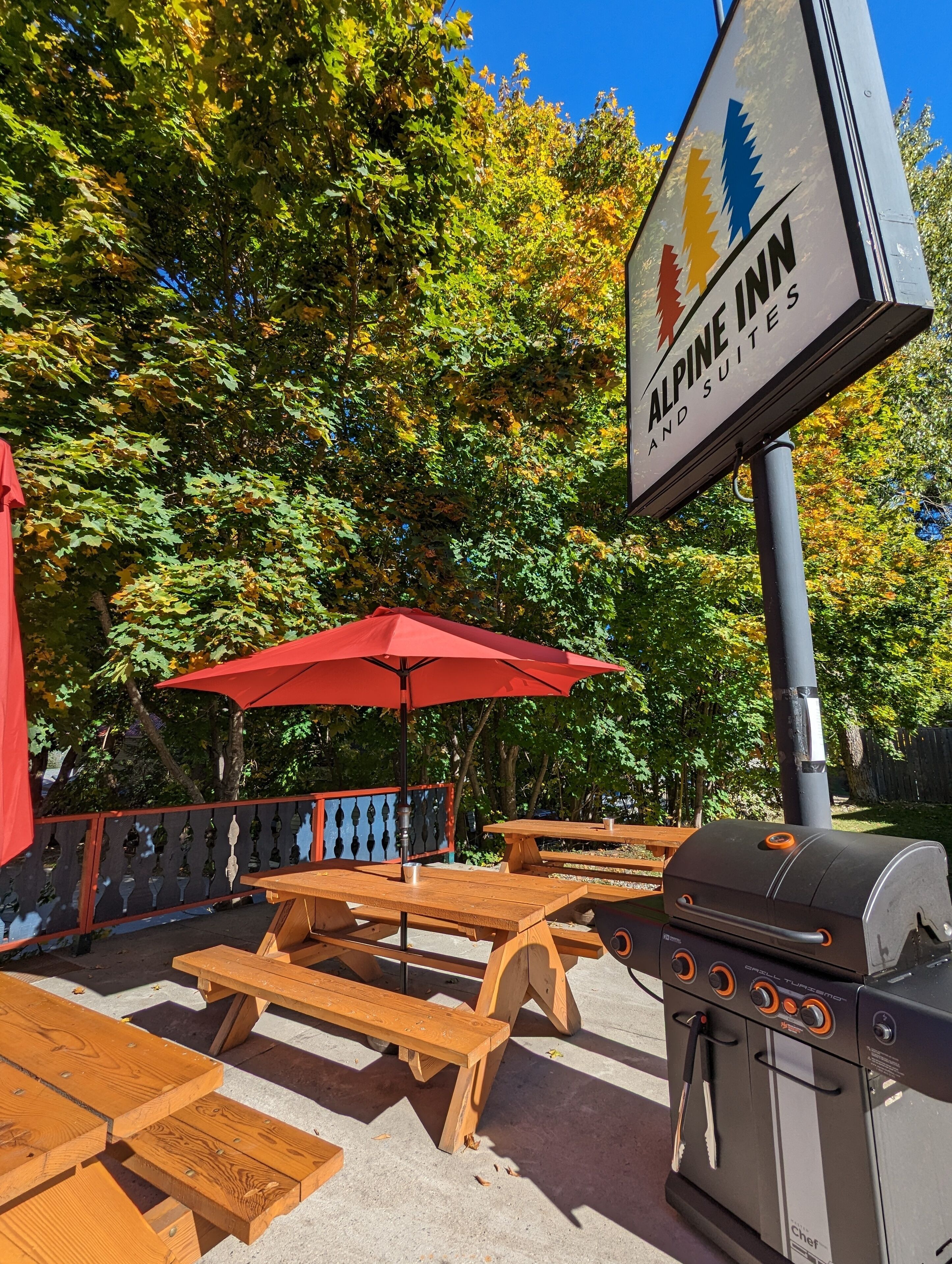 Een terras buiten met houten tafels en rode parasols, een barbecue en een bord met Alpine Inn and Suites.