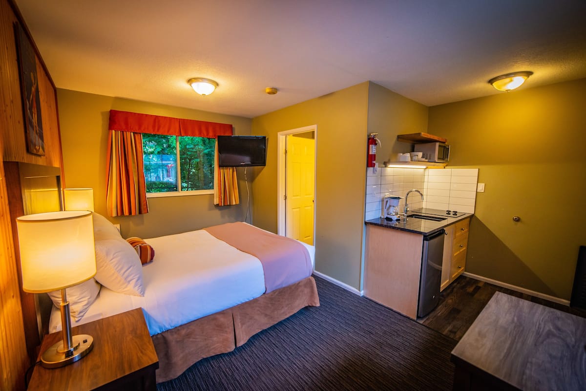 A hotel room with a bed, bedside table, lamp, TV, and a kitchenette with a microwave and sink.