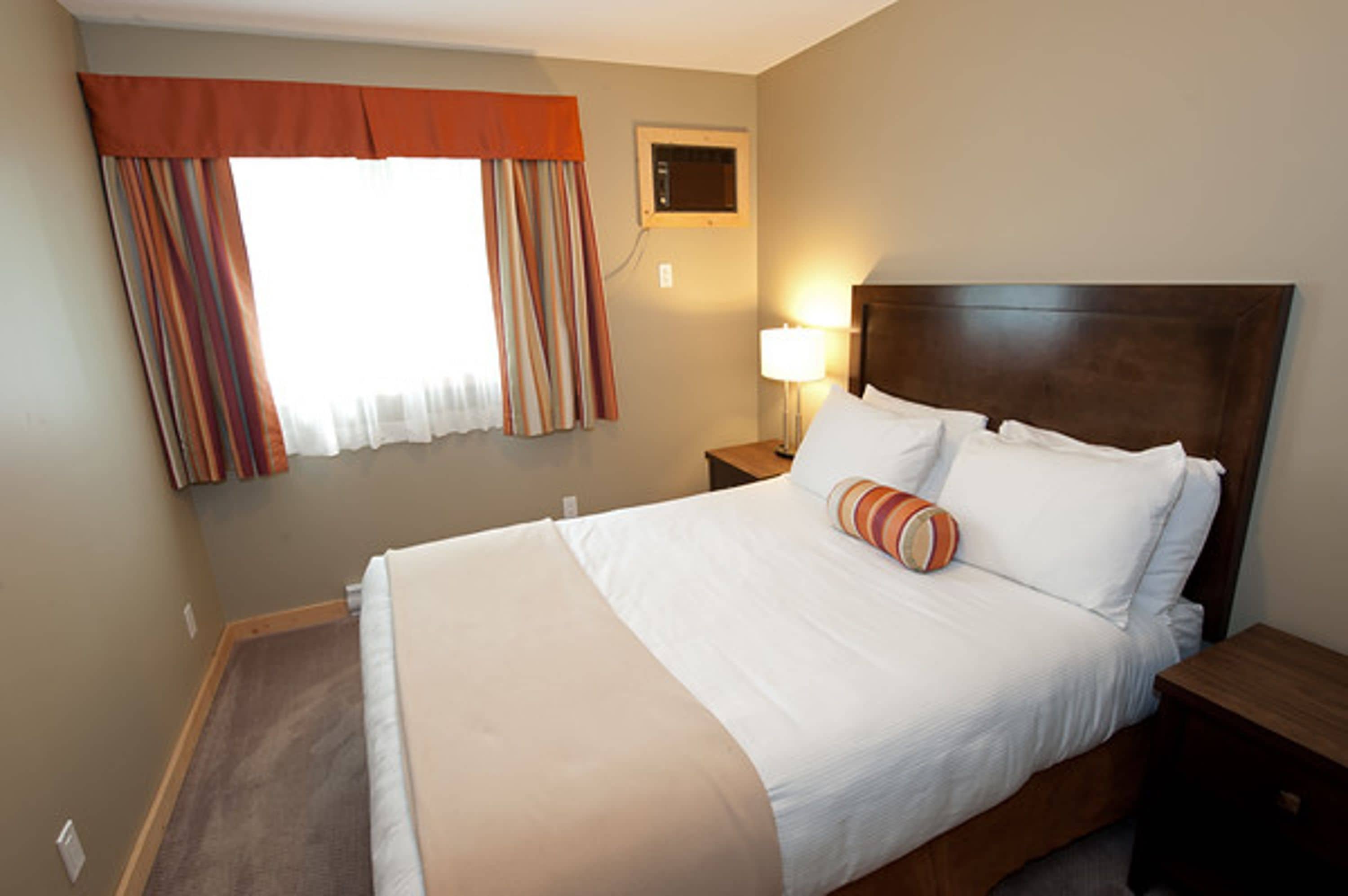Een hotelkamer met een groot bed, een nachtkastje met een lamp, een raam met gordijnen en een wandairconditioning.