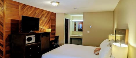 A hotel room with a wooden wall panel design, a bed with white bedding, a television, a microwave, a desk with a lamp, and a bathroom visible through an open door.