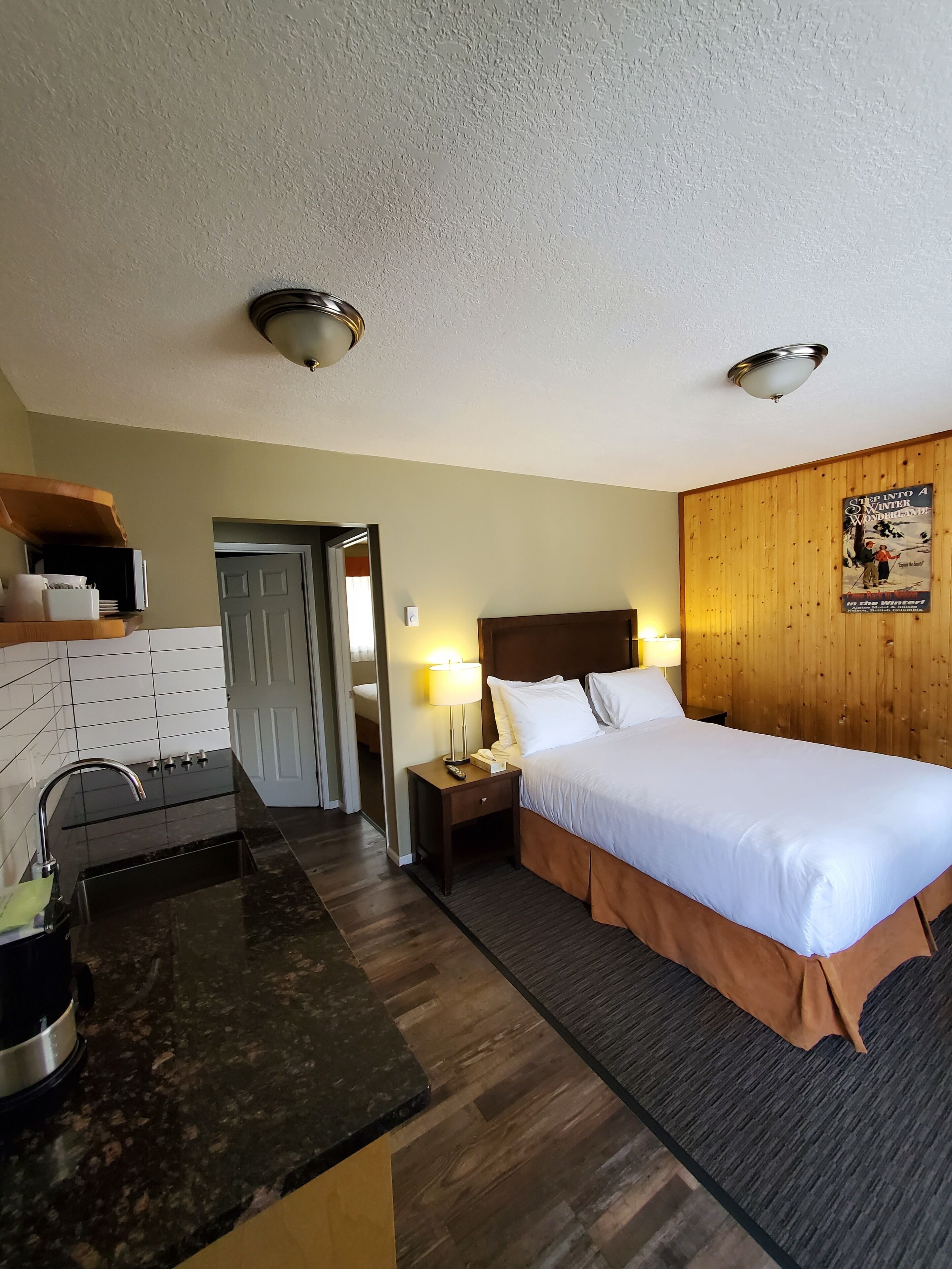 A hotel room with a bed, a nightstand, a lamp, a wooden headboard, a wooden wall panel, a door, and a kitchen area with a sink and countertop.