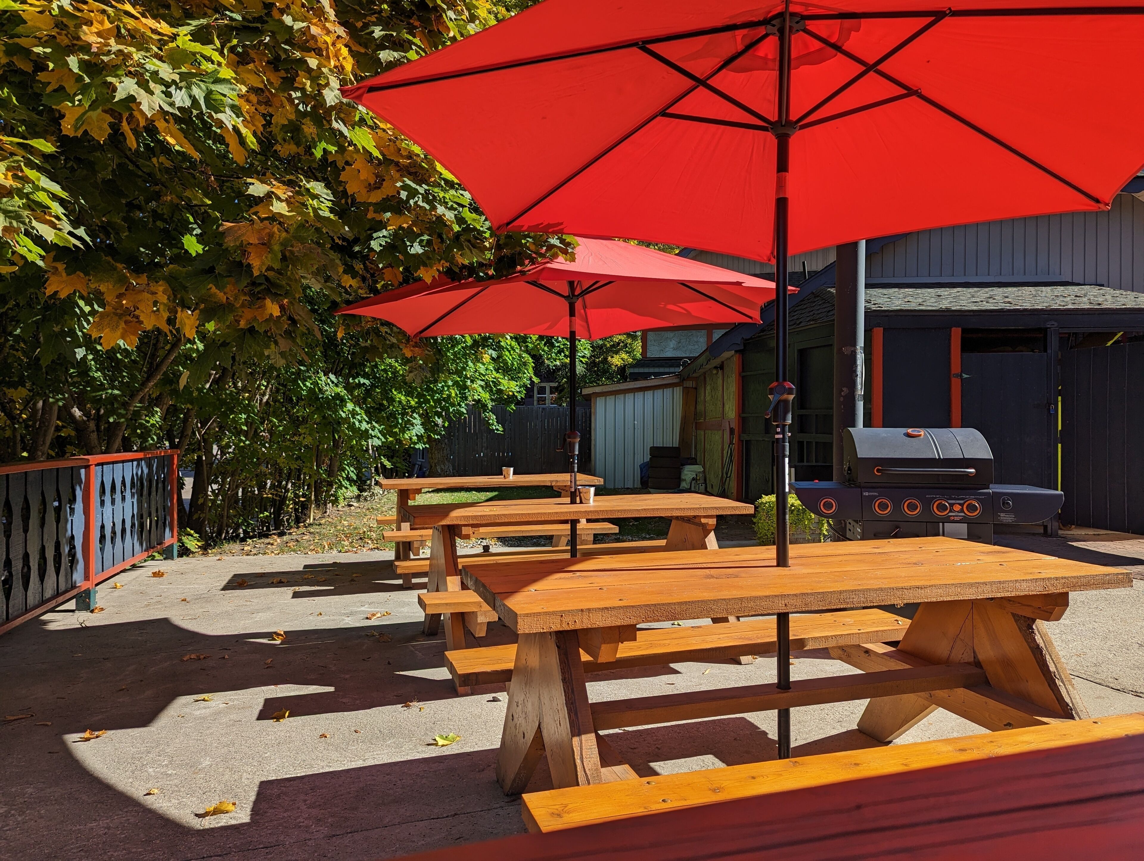 Outdoor wooden picnic tables with red umbrellas, a barbecue grill, and a building in the background.