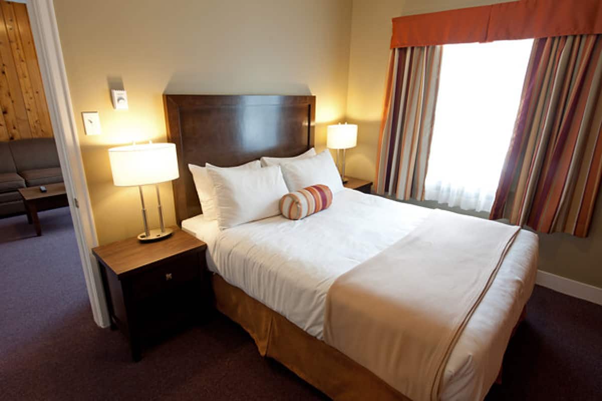 A neatly made bed with white linens and a striped pillow, a wooden nightstand with a lamp, and a window with curtains.