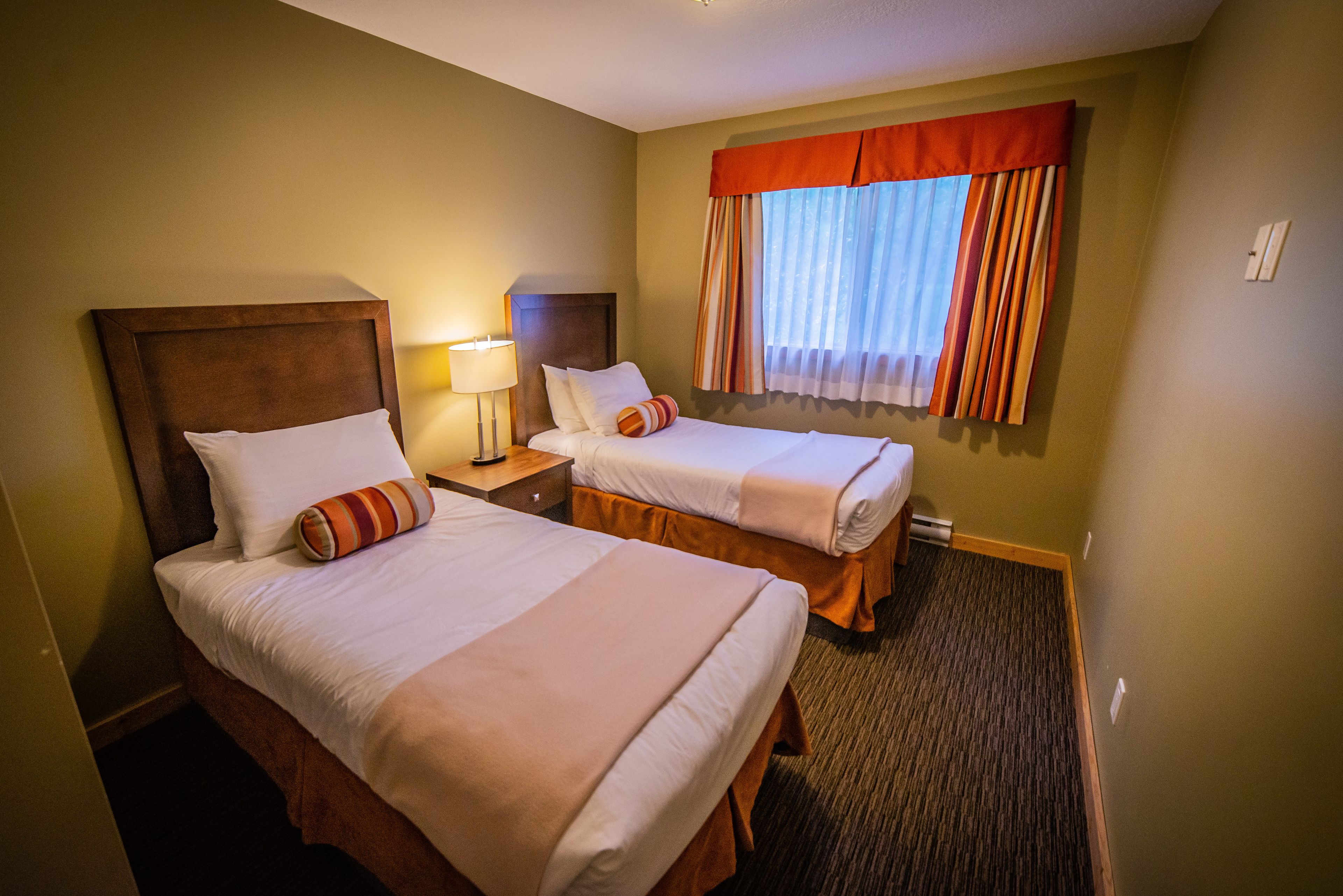 A hotel room with two beds, a nightstand, a lamp, and a window with curtains.