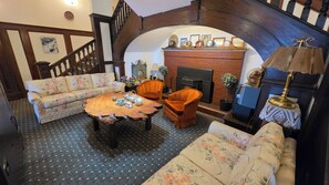 A cozy living room with a fireplace, floral-patterned sofas, a wooden coffee table, and a staircase. - Rodmay Heritage Hotel (Powell River)