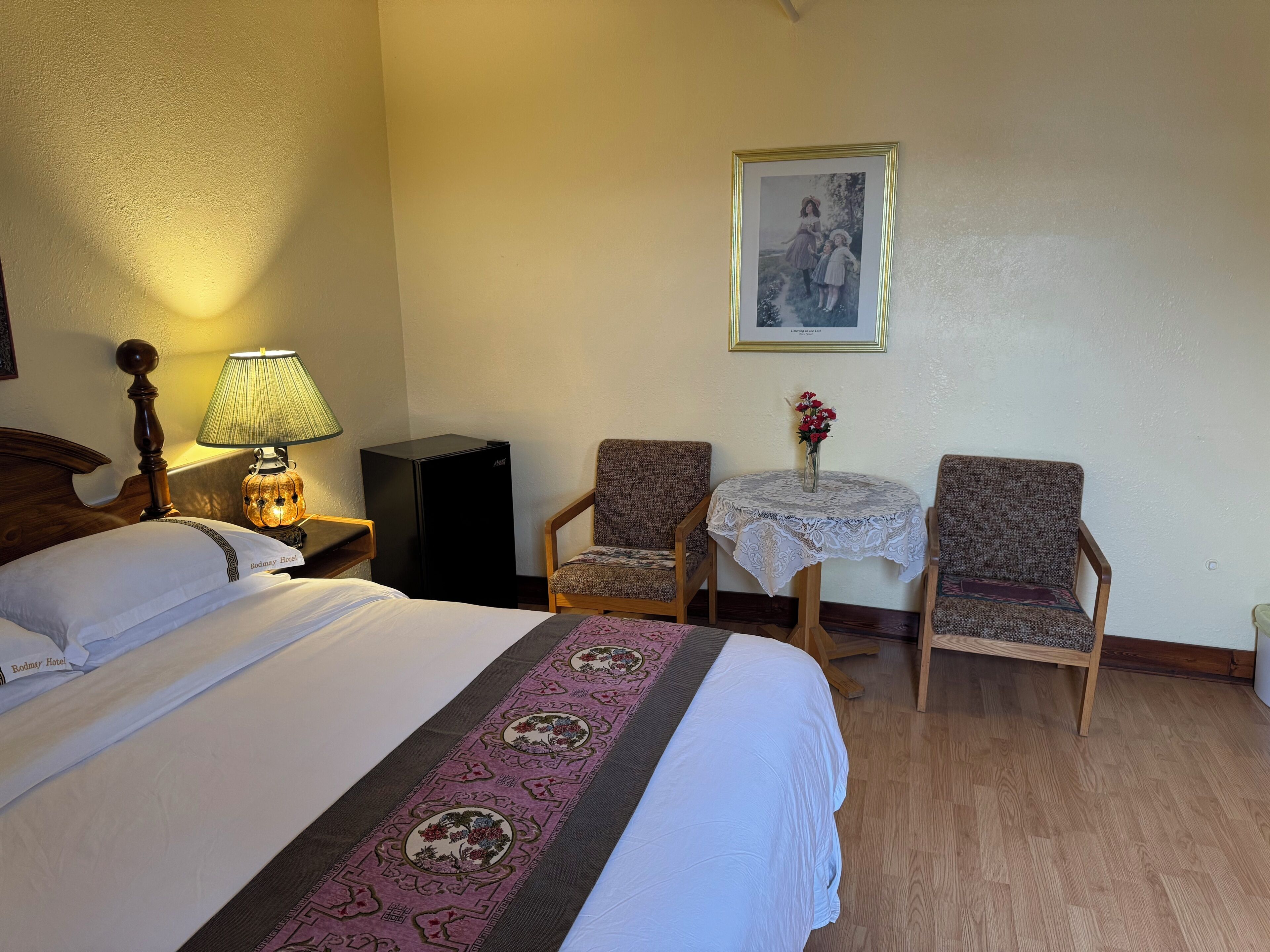Habitación de hotel con cama, mesita de noche con lámpara, pequeña mesa con un jarrón de flores, dos sillones y un cuadro en la pared.