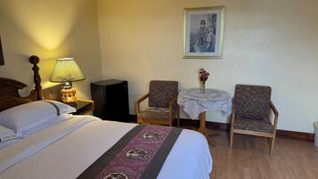 Habitación de hotel con cama, mesita de noche con lámpara, pequeña mesa con un jarrón de flores, dos sillones y un cuadro en la pared.