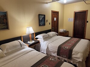 A hotel room with two beds, a nightstand with a lamp, a framed picture on the wall, and a door with a sign. - Rodmay Heritage Hotel (Powell River)