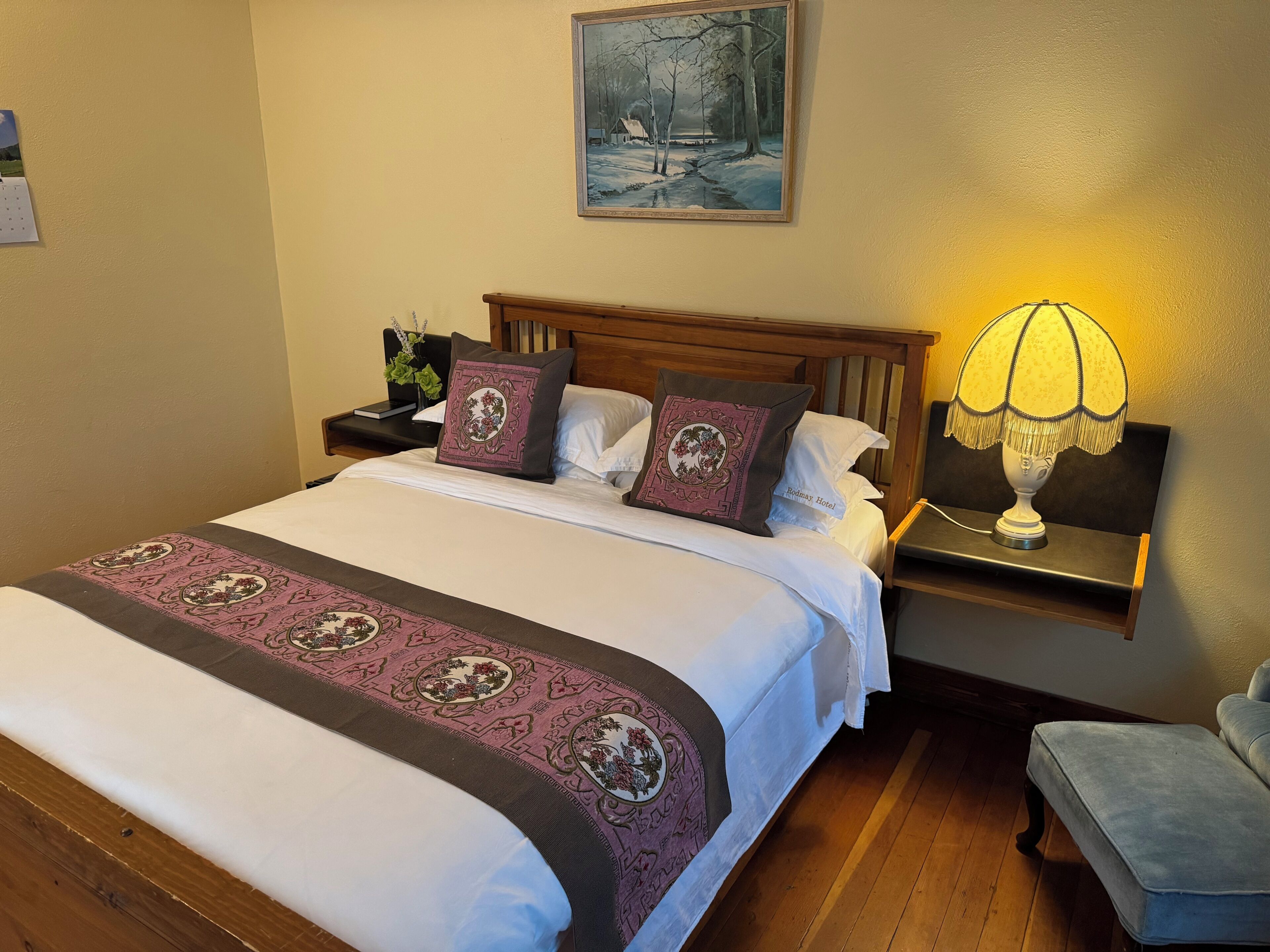 Una cama bien hecha con un edredón estampado, una mesita de noche con lámpara, un cuadro en la pared y una silla en la esquina.