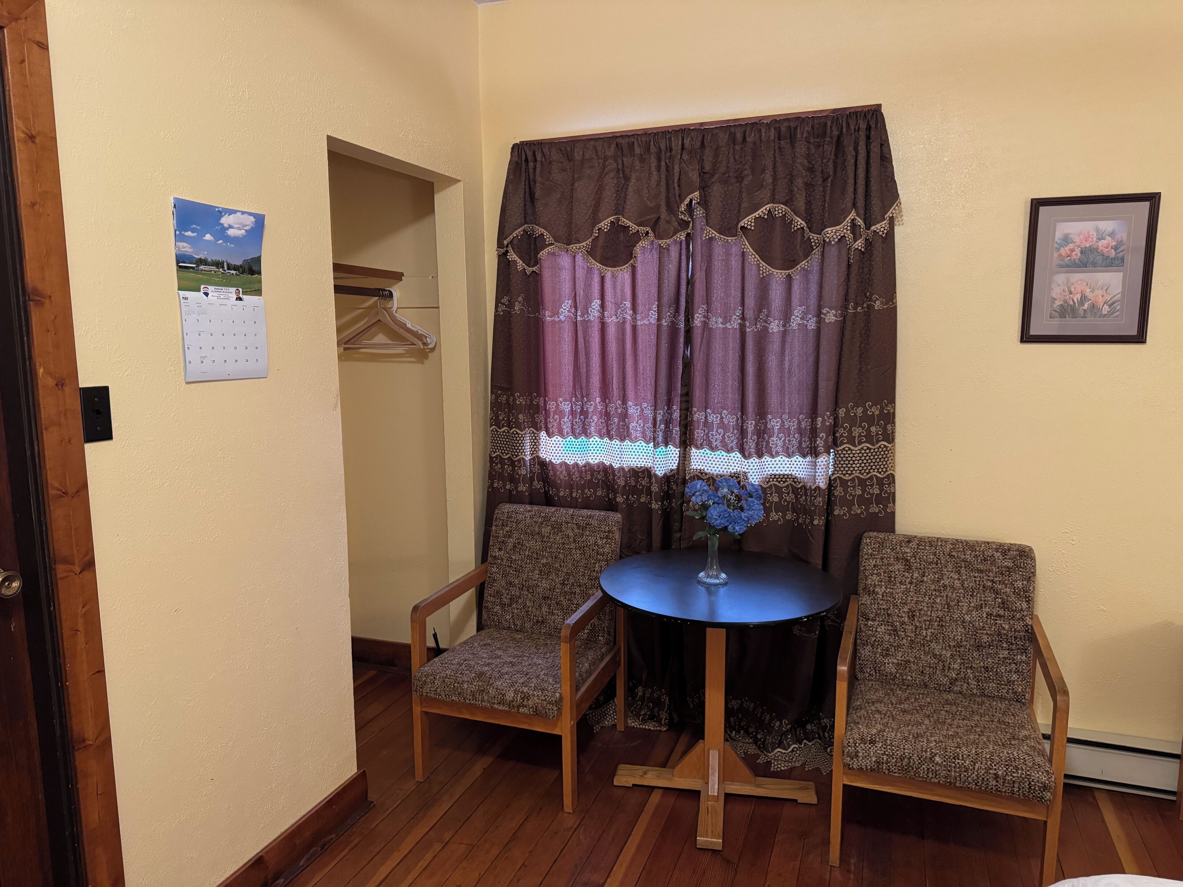 A small room with two chairs, a table, a window with curtains, a calendar, and a framed picture on the wall.