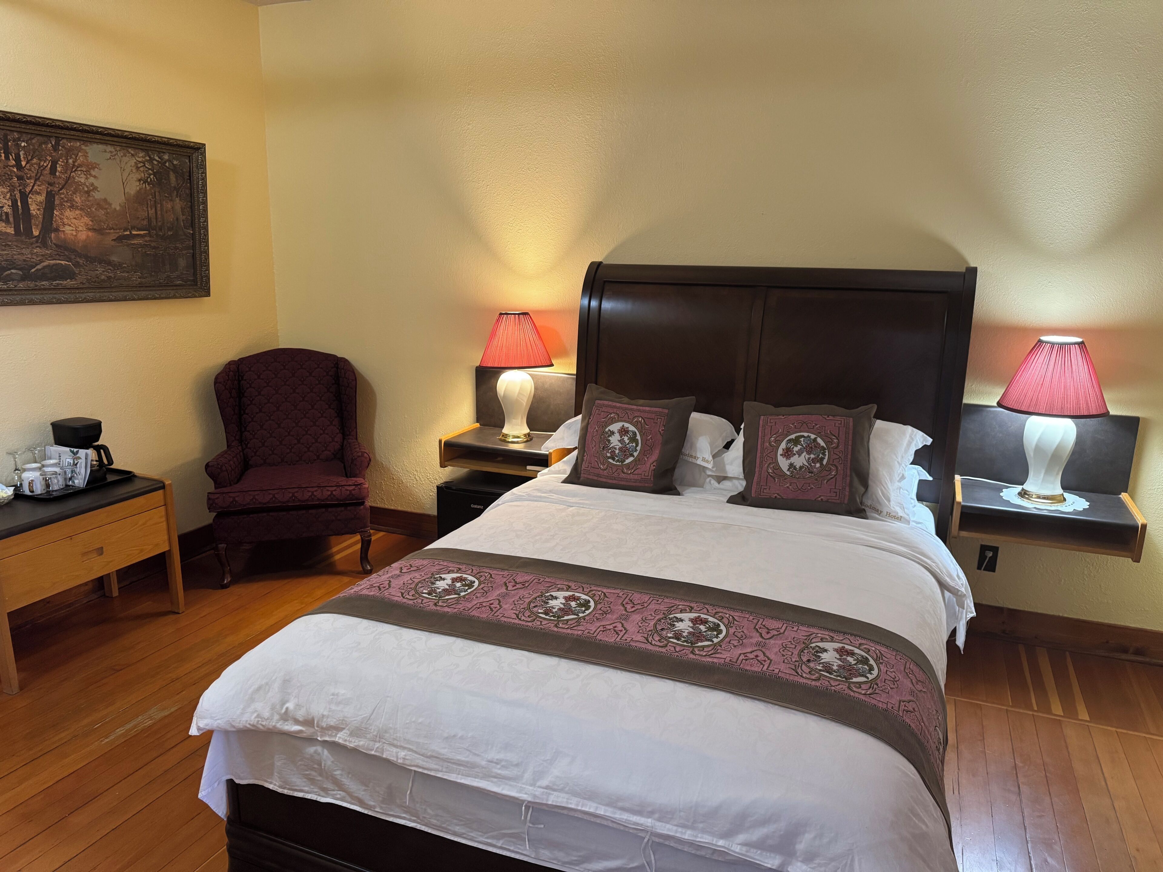 Una habitación de hotel con cama, dos mesitas de noche, una silla, un cuadro en la pared y una zona de café.