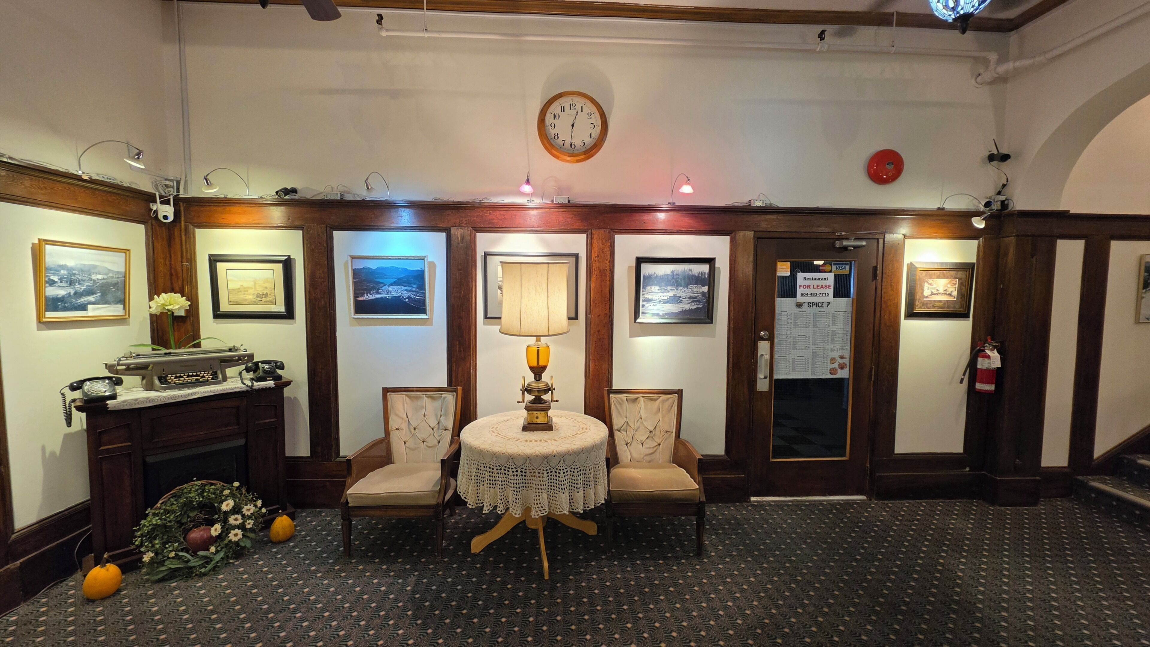 A hotel lobby with a round table, chairs, framed pictures, a clock, and a door with a sign.