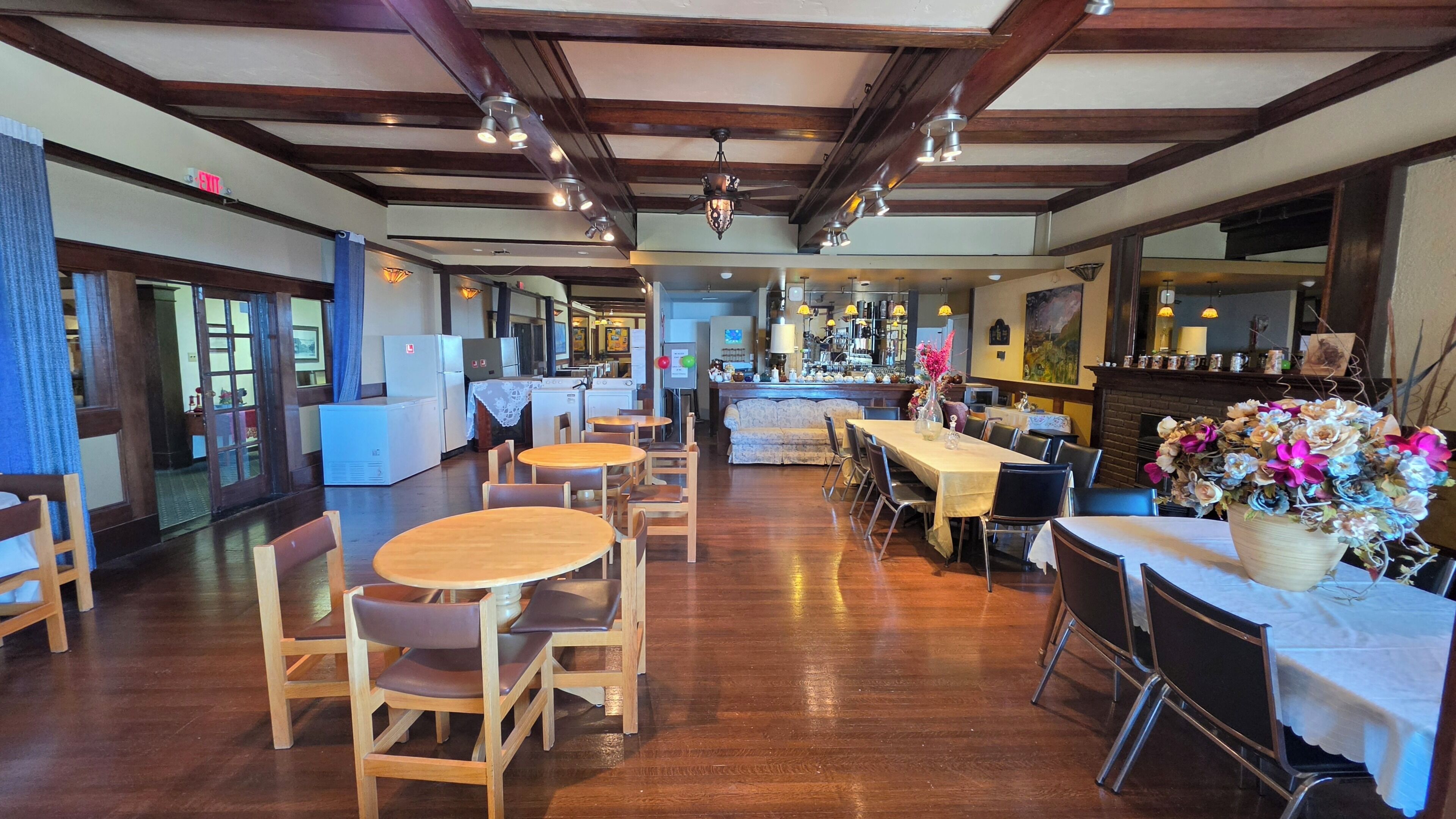 A spacious restaurant with wooden tables and chairs, a bar area, and a view into the kitchen.
