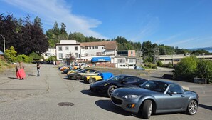A parking lot with several sports cars, a building with a blue awning, and a view of trees and hills in the background. - Rodmay Heritage Hotel (Powell River)