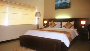 Une chambre d’hôtel avec un grand lit, une table de chevet, une lampe, un tableau au mur et une fenêtre avec des stores.
