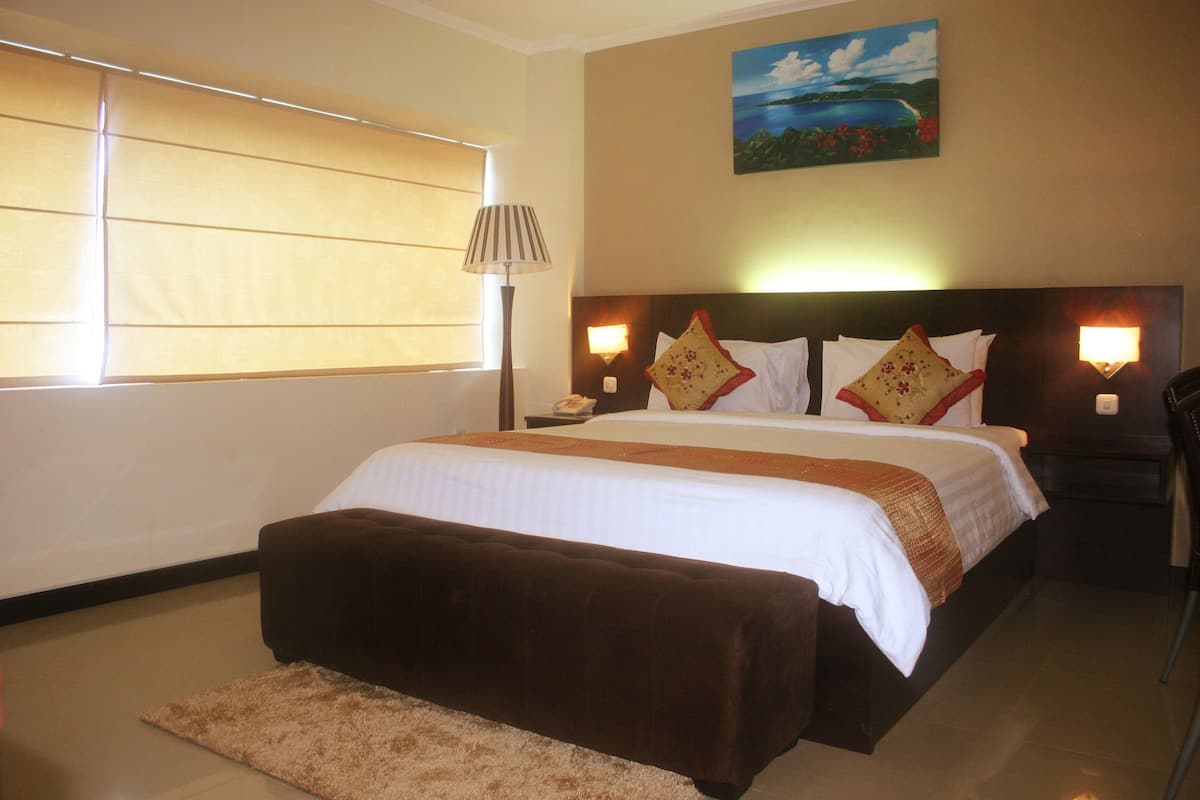 Habitación de hotel con una cama grande, una mesita de noche, una lámpara, un cuadro en la pared y una ventana con persianas.