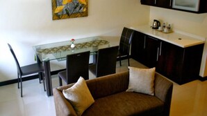 Un salon avec un canapé marron, deux coussins, une table à manger en verre avec des chaises, un coin cuisine avec des placards et un tableau accroché au mur.