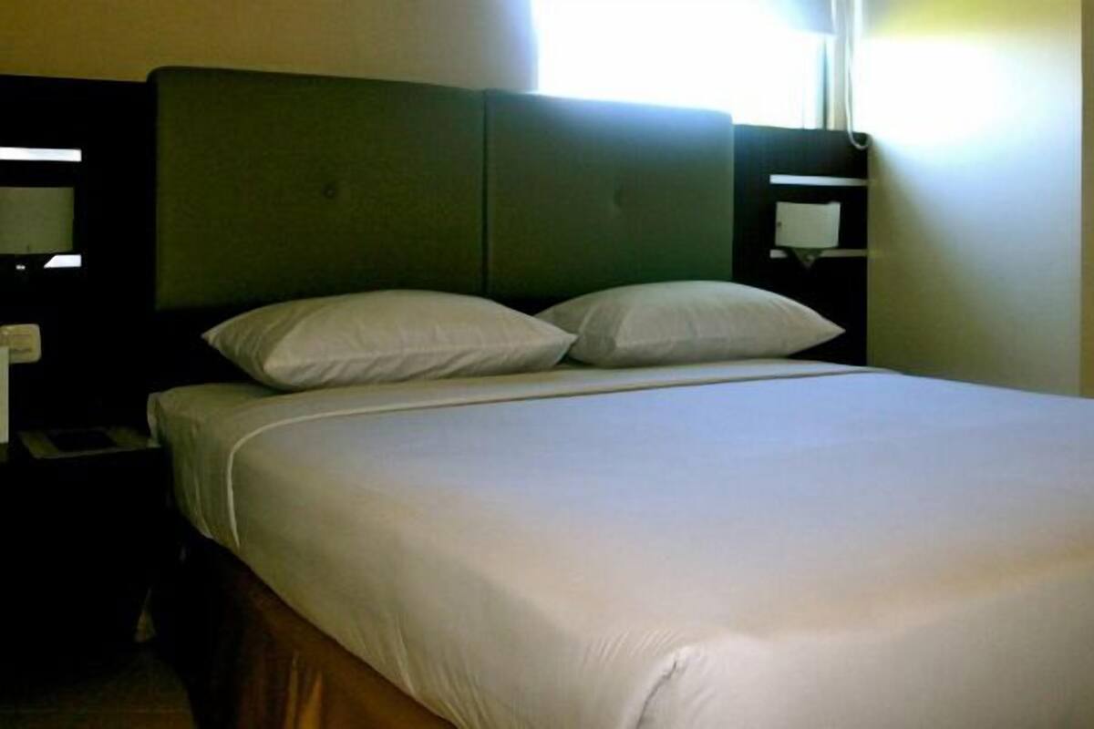 Una cama bien tendida con un cabecero verde, almohadas blancas y una mesita de noche de madera con una lámpara.