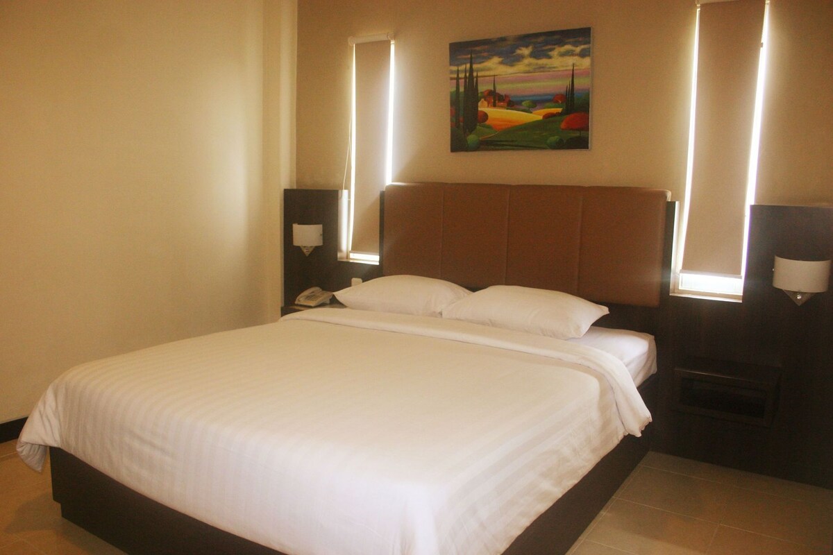 Un cuarto de hotel con una cama grande, lámparas de noche, un cuadro en la pared y una mesita de noche con un teléfono.