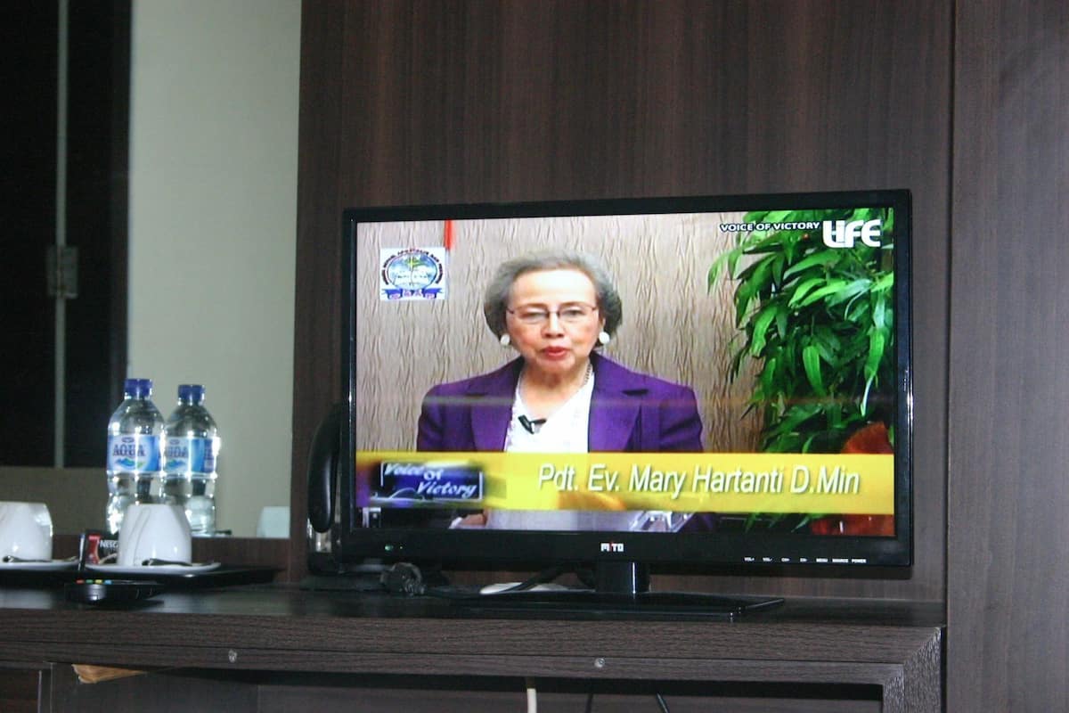 Un televisor mostrando un programa de noticias con una mujer con un blazer púrpura, una planta y botellas de agua sobre una mesa.