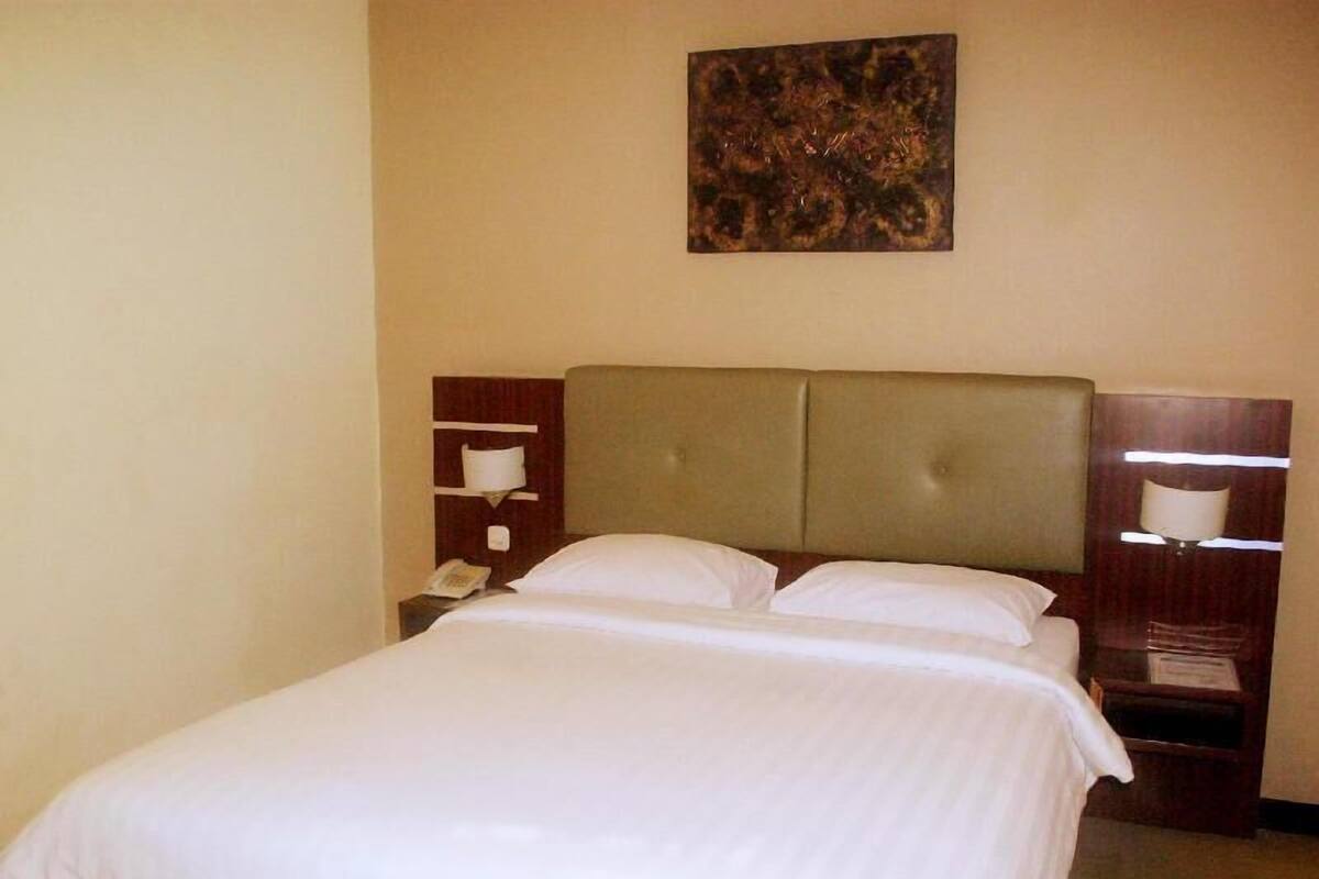 Habitación de hotel con cama, cabecero, mesita de noche con lámpara y una obra de arte colgada en la pared.