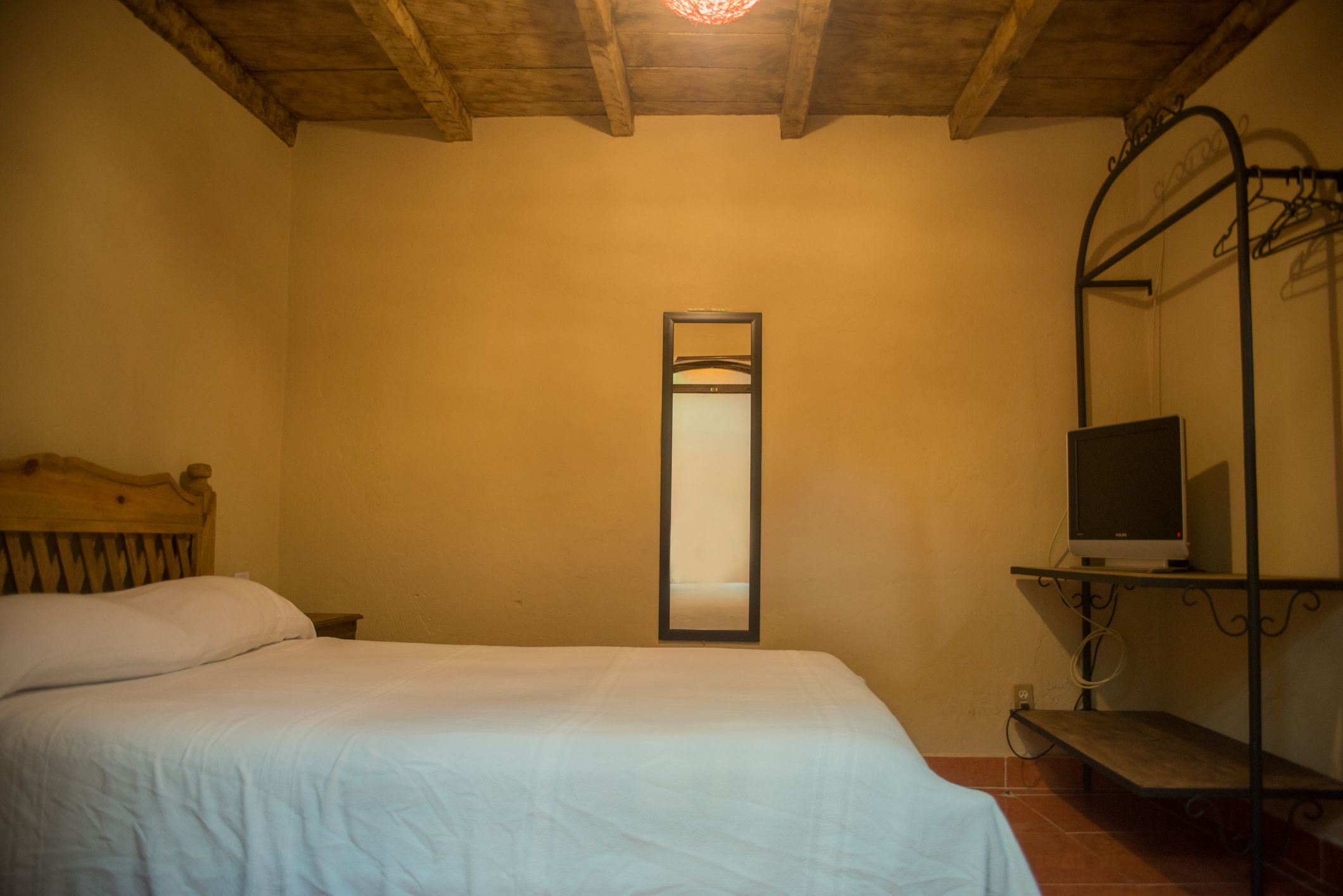 Un dormitorio con techo de madera, una cama con ropa de cama blanca, una mesita de noche, un espejo y un televisor sobre una estantería.