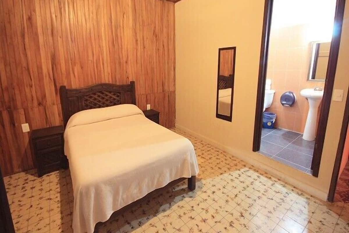 Une chambre à coucher avec une tête de lit en bois, un lit simple, une table de chevet, un miroir et une salle de bain visible à travers une porte ouverte.