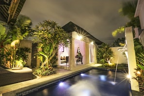 A swimming pool with a glass enclosure, surrounded by tropical plants and evening lighting.