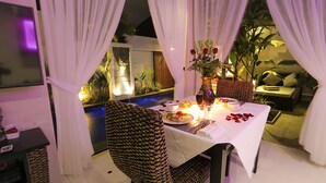 Un comedor con una mesa dispuesta para dos personas, rodeado de cortinas blancas, un jarrón con flores y vistas a una piscina y plantas.