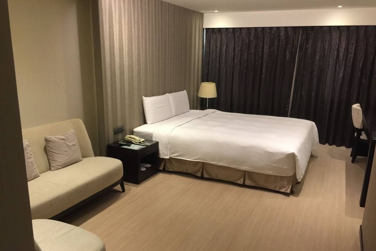 A hotel room with a bed, a sofa, a nightstand with a lamp, and a chair.