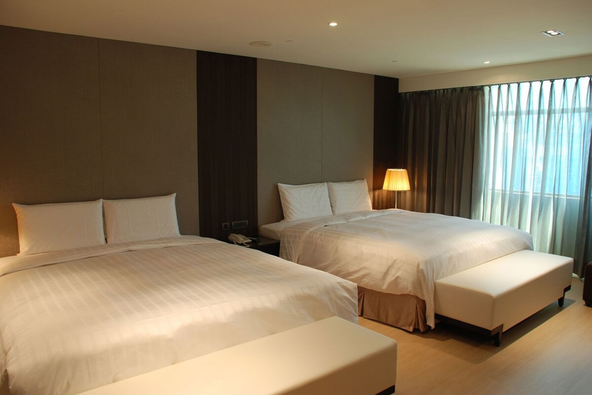 Una habitació d'hotel amb dos llits, una taula de nit amb una làmpada i una finestra gran amb cortines.