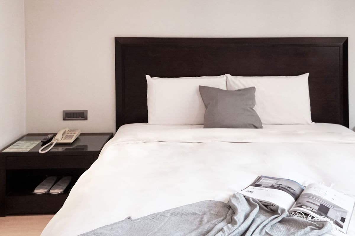 A hotel room with a bed, a nightstand with a telephone, and a magazine on the floor.